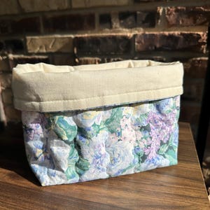 May include: A rectangular fabric basket with a floral pattern in shades of blue, green, and purple. The top edge is a solid cream color, and the basket appears to be quilted. The basket is sitting on a wooden surface.