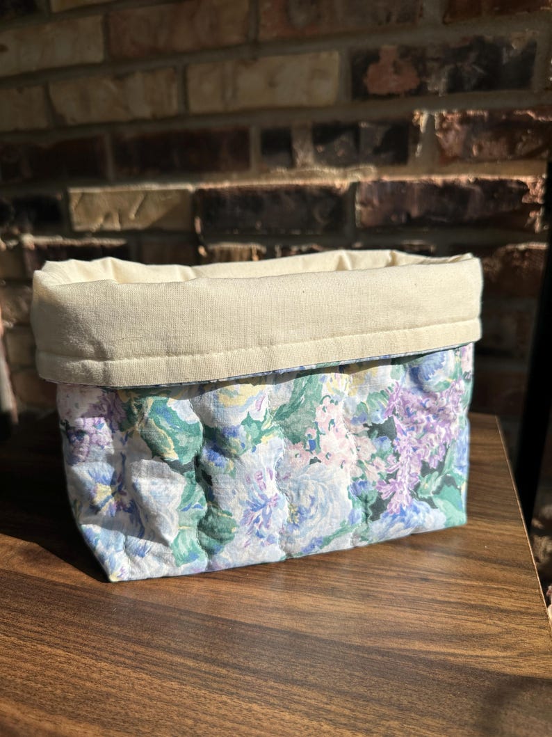 May include: A rectangular fabric basket with a floral pattern in shades of blue, green, and purple. The top edge is a solid cream color, and the basket appears to be quilted. The basket is sitting on a wooden surface.