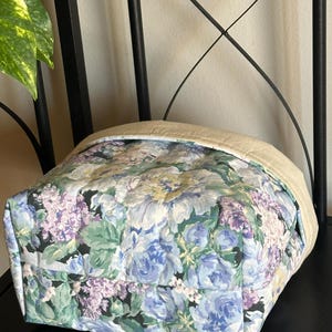 May include: A quilted fabric tote bag with a floral pattern in shades of blue, purple, and green. The top edge is a solid beige color. The bag is sitting on a black surface.