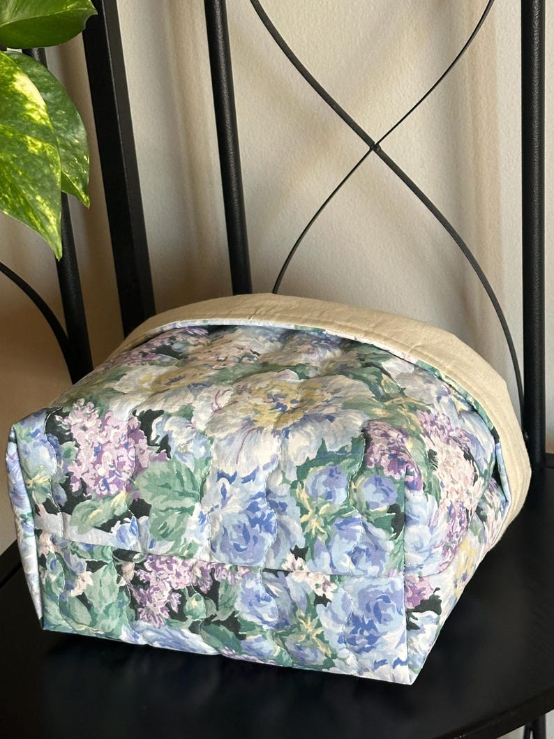 May include: A quilted fabric tote bag with a floral pattern in shades of blue, purple, and green. The top edge is a solid beige color. The bag is sitting on a black surface.