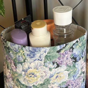 May include: A fabric storage bin with a floral pattern in shades of blue, purple, and green. The bin contains various toiletries, including bottles and tubes. The bin is lined with a neutral fabric.
