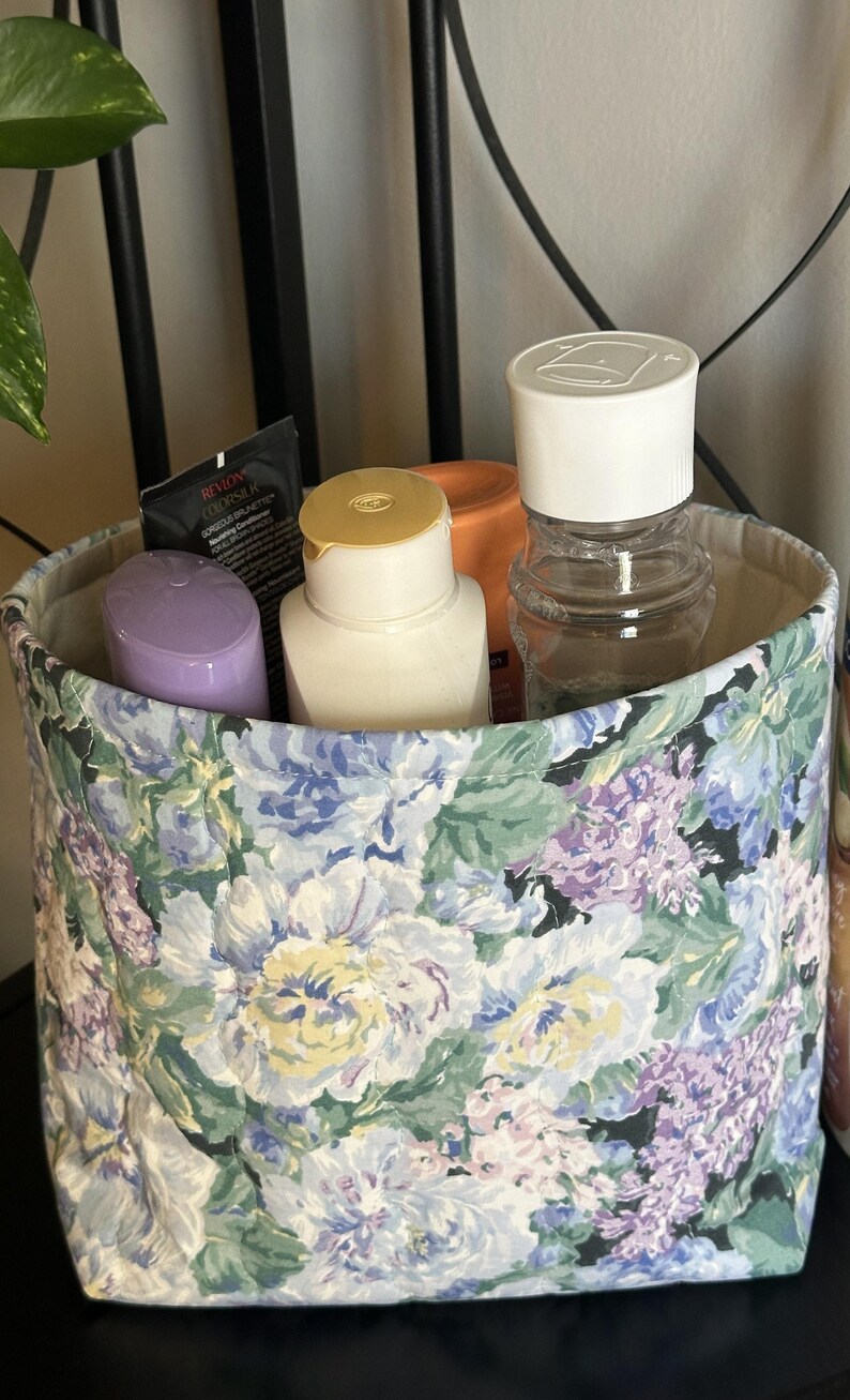 May include: A fabric storage bin with a floral pattern in shades of blue, purple, and green. The bin contains various toiletries, including bottles and tubes. The bin is lined with a neutral fabric.