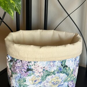 May include: A fabric storage basket with a floral pattern in shades of blue, purple, and yellow. The top edge is a solid beige color. The basket is sitting on a black surface.