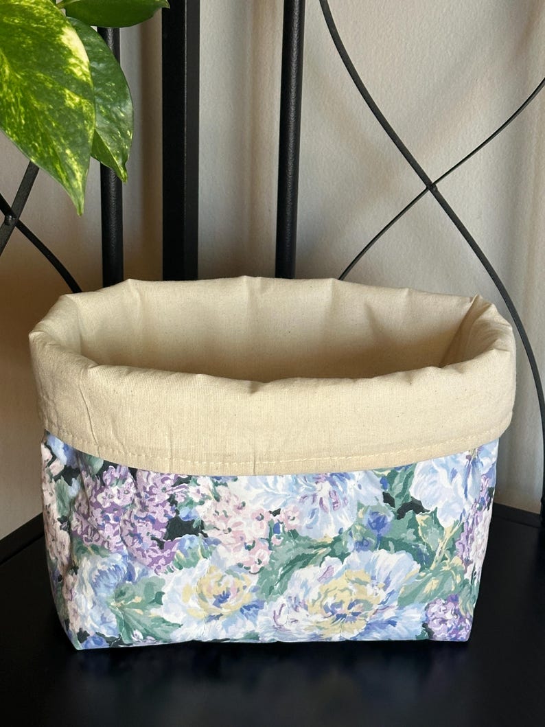 May include: A fabric storage basket with a floral pattern in shades of blue, purple, and yellow. The top edge is a solid beige color. The basket is sitting on a black surface.