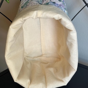 May include: A fabric storage basket with a floral pattern on the upper section. The basket's interior and lower exterior are a natural cream color. The floral pattern includes shades of blue, green, and purple.