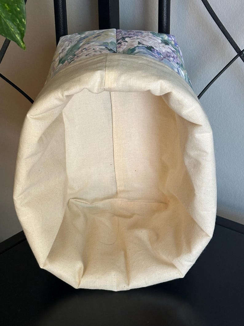 May include: A fabric storage basket with a floral pattern on the upper section. The basket's interior and lower exterior are a natural cream color. The floral pattern includes shades of blue, green, and purple.