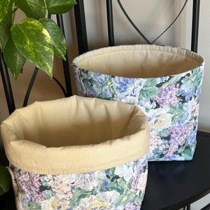 May include: Two fabric storage baskets. The exterior features a floral pattern in shades of blue, purple, and green on a black background. The interior is a solid beige color. The baskets are different sizes and are sitting on a black surface.