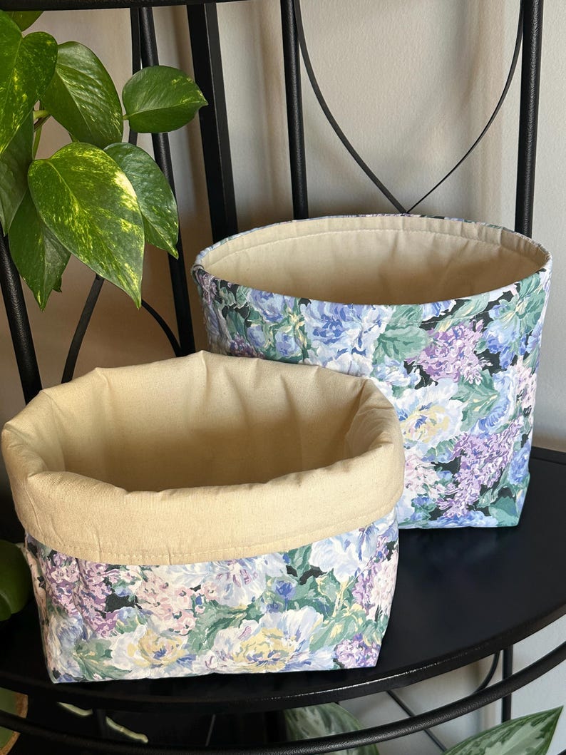 May include: Two fabric storage baskets. The exterior features a floral pattern in shades of blue, purple, and green on a black background. The interior is a solid beige color. The baskets are different sizes and are sitting on a black surface.