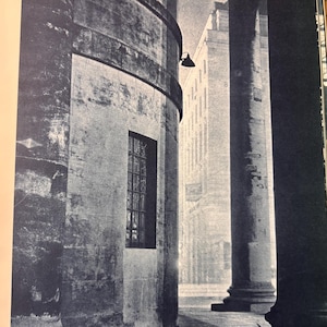 May include: Monochrome photograph of a building with a curved wall and a small, gridded window. Large columns are in the foreground, with another building visible in the background. The image is in black and white.