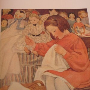 May include: A young girl in a red dress sits in a chair and sews a white fabric. She is surrounded by dolls, some of which are wearing striped dresses.