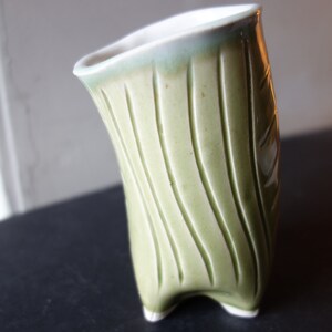 Op de afbeelding: Een handgemaakte, lichtgroene keramische vaas met een unieke, gebogen vorm. De vaas heeft verticale, getextureerde lijnen en een wit interieur. Hij staat op drie kleine voetjes en heeft een lichtgroene rand.