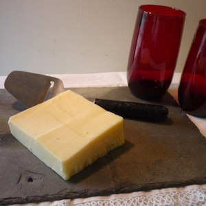 Puede incluir: Un bloque de queso amarillo pálido sobre una tabla de servir de pizarra con un rallador de queso y un cuchillo de mango negro. Dos vasos rojos de vidrio están en el fondo.