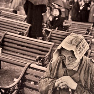 Può includere: Fotografia in bianco e nero di una giornata piovosa. Una persona anziana è seduta su una panchina, riparandosi con un giornale a mo' di cappello. Altre persone, alcune con ombrelli, sono visibili. L'immagine ha un'atmosfera vintage.