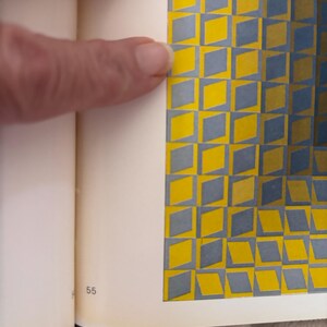 May include: A book page with a geometric pattern of yellow and gray diamonds. The page is open, with a finger pointing at the pattern. The number 55 is visible at the bottom of the page.
