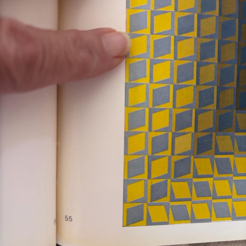 May include: A book page with a geometric pattern of yellow and gray diamonds. The page is open, with a finger pointing at the pattern. The number 55 is visible at the bottom of the page.