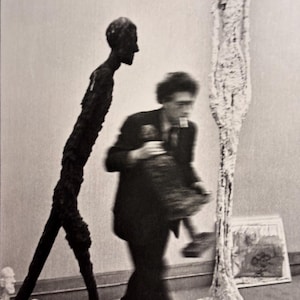 Könnte beinhalten: Schwarz-Weiß-Fotografie eines Künstlers in einem Atelier mit Skulpturen. Eine hohe, dünne, dunkle Skulptur einer Figur steht in der Nähe einer verschwommenen Figur eines Mannes, der eine Skulptur hält. Eine weiße Skulptur und ein Gemälde sind ebenfalls zu sehen.