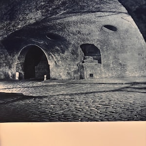 May include: A black and white photo of a brick tunnel with a cobblestone floor. The tunnel has a vaulted ceiling with a small round opening in the ceiling. There are two arched doorways on the left and right sides of the tunnel.