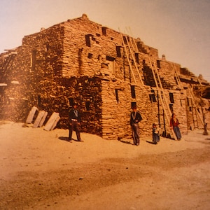 May include: A large, multi-story stone building with wooden ladders leaning against the exterior. The building is made of rough-hewn stones and has a rustic, weathered appearance. There are several people standing in front of the building, including a man in a cowboy hat and a woman in a long dress.