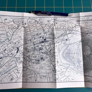 May include: Folded vintage map of Edinburgh, Scotland, with detailed street layouts and landmarks. The map is printed in black and white with a blue pen resting on top. The title "Plan of Edinburgh" is visible.