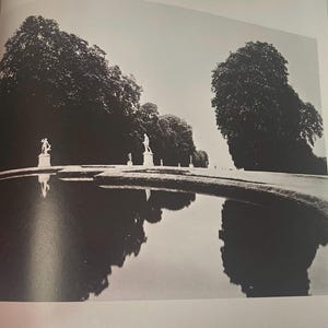 Puede incluir: Fotografía en blanco y negro de un jardín formal con un estanque reflectante. Estatuas se colocan a lo largo del borde del estanque y a lo lejos. Grandes árboles enmarcan la escena, con sus reflejos reflejados en el agua.