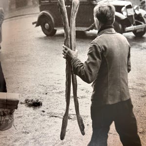 Può includere: Un uomo con una giacca e pantaloni scuri cammina per strada portando due lunghi bastoni di legno. Un'auto d'epoca è parcheggiata sullo sfondo.