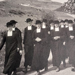 Può includere: Fotografia in bianco e nero di un gruppo di persone che indossano abiti neri, cappelli e colletti bianchi, che camminano su un sentiero. L'immagine è scattata all'aperto, con un paesaggio collinare sullo sfondo.
