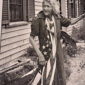 May include: A black and white photograph of an elderly person wearing a dress and holding an American flag. The flag is draped around the person's neck and chest. The person is standing in front of a weathered building.