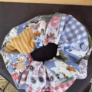 May include: A fabric hair scrunchie featuring a patchwork design of various patterns including blue and white gingham, yellow stripes, and multiple floral prints in shades of pink, blue, green, and white, gathered into a circular shape.