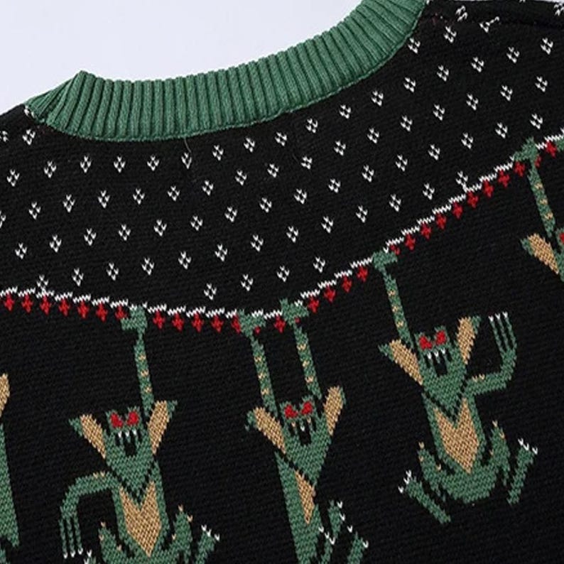 May include: Black knitted sweater with a green trim around the neck. The sweater has a repeating pattern of green, beige, and red creatures with red eyes hanging from a line.