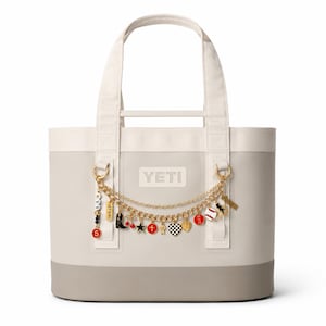 Könnte beinhalten: Eine zweifarbige Tragetasche mit cremefarbenem Oberteil und grauem Boden. Die Tasche hat einen cremefarbenen Griff und eine goldene Kette mit verschiedenen Anhängern. Das Wort "YETI" ist auf der Vorderseite der Tasche aufgedruckt.