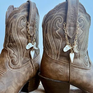 May include: A pair of brown leather Durango cowboy boots featuring intricate tonal stitching and a gold-toned chain accessory. The chain is adorned with small white enamel charms shaped like a cowboy hat, a boot, and a heart with a star detail.