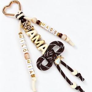 May include: A keychain featuring a gold heart-shaped clasp, cream-colored cord, and brown rope. It includes beads spelling MAMA, MAGS, and MILO MIA, with a small heart charm and a brown knotted rope accent with cream-colored heart-shaped beads.