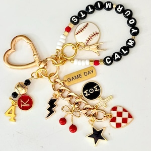 May include: A gold-toned keychain with various charms, including a baseball, a lightning bolt, a cross, and a heart. The keychain also features a charm with the text "GAME DAY" and a beaded bracelet with the words "CALM YOUR TITS".