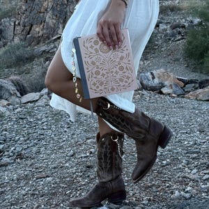 May include: A light pink notebook with a dark gray spine and a white floral pattern on the cover, featuring a dangling charm accessory with colorful beads and metal icons, paired with dark brown leather cowboy boots and a white skirt.