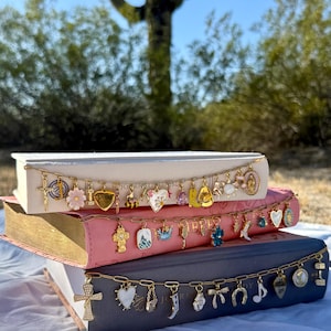 May include: Three stacked books with gold-edged pages, each adorned with a gold chain of charms. The charms include crosses, hearts, flowers, and other religious and decorative elements. The books are in shades of white, pink, and blue.