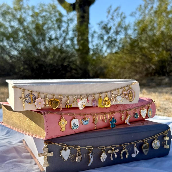 May include: Three stacked books with gold-edged pages, each adorned with a gold chain of charms. The charms include crosses, hearts, flowers, and other religious and decorative elements. The books are in shades of white, pink, and blue.