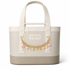 Könnte beinhalten: Eine hellbeige YETI-Tragetasche mit einer goldfarbenen Kette, die über die Vorderseite drapiert ist. Die Kette ist mit verschiedenen kleinen Anhängern verziert, darunter ein rosa Stern, ein Herz, ein Marienkäfer, eine Schleife, eine Tänzerin, eine Blume, ein Kreuz und eine kleine rosa Tasche.