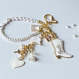May include: A gold-toned keychain with multiple charms, including a white cowboy boot with "Bride" written on it, a heart, a cross, and a diamond. The keychain also features a pearl beaded strand with the word "TROPHY".