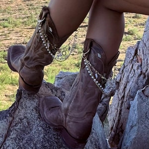 Puede incluir: Botas de vaquero de cuero marrón con cadenas y abalorios decorativos. Las botas tienen un diseño occidental clásico con costuras intrincadas y una puntera cuadrada. Las cadenas están adornadas con perlas y otros adornos.