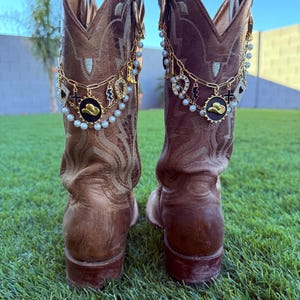 May include: Two brown leather cowboy boots, adorned with gold chains, charms, and faux pearls. Charms include a horseshoe, cross, and cowboy hat. The boots are displayed on a green grassy surface.