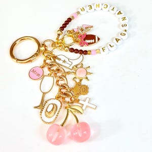 May include: A gold-toned keychain with various charms, including a football, cowboy boot, sun, cross, and cherries. The keychain also features a white beaded bracelet with the text "FLAG ATTACHER". Additional charms include a wine glass and a pink circle with the word "Mom".