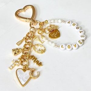 May include: A gold-toned keychain featuring a heart-shaped clasp, a circular strand of white beads spelling LOVE STORY, and various gold charms including a cross, a heart, a key, a moon, and the script word mrs, with a small heart charm engraved love you.