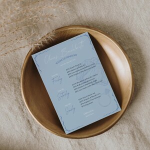 May include: A light blue card with a white outline and a floral design. The card lists a weekend itinerary for a bachelorette party with the days of the week, times, and activities. The text on the card reads "Olmaz's Bachelorette Weekend Itinerary".