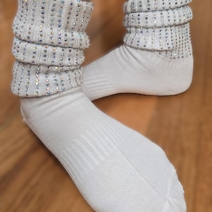 May include: A pair of white socks with a scrunched, embellished cuff. The cuffs are adorned with rows of iridescent rhinestones. The socks are made of a soft, ribbed material, perfect for adding a touch of sparkle to any outfit.