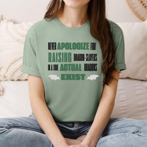 Könnte beinhalten: Salbeigrünes T-Shirt mit dem Text "ENTSCHULDIGEN SIE SICH NIEMALS DAFÜR, DRACHENTÖTER AUFZUZIEHEN, IN EINER ZEIT, IN DER TATSÄCHLICH DRACHEN EXISTIEREN" in Schwarz und Grün. Das Shirt hat eine Fledermausflügel-Grafik. Das Shirt ist aus weichem Material.
