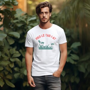 May include: White t-shirt with the word "PRO-LE-TAR-I-AT" in pink letters. Below the text is a teal graphic of a crowd of people holding flags. The shirt is being worn by a person with dark hair and is paired with gray pants.