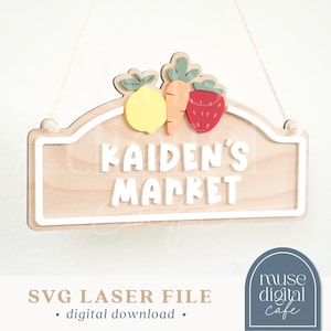 Puede incluir: Letrero de madera con el texto "KAIDEN'S MARKET" en blanco, coronado por un limón, una zanahoria y una fresa. El letrero es de color marrón claro con un borde blanco y cuelga de una cuerda blanca. Las palabras "SVG LASER FILE" y "digital download" están en la parte inferior.