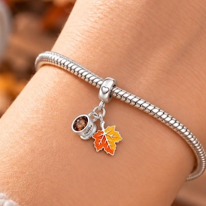 Può includere: Un bracciale a catena snake in argento con un set di ciondoli pendenti che include una piccola tazza da caffè bianca con liquido marrone e una foglia d'acero dai colori arancione e giallo. Il ciondolo è fissato da una piccola perlina argentata a cuore.