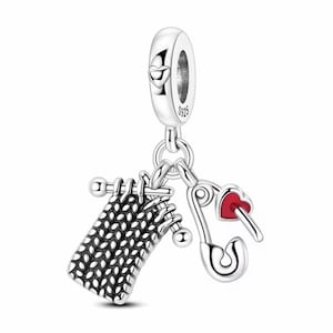 Könnte beinhalten: Ein silberner Charm mit Strickmotiv. Es zeigt ein Strickstück mit Nadeln, eine Sicherheitsnadel mit rotem Herz und einen herzförmigen Bügel. Der Charm ist für ein Armband oder eine Halskette konzipiert.