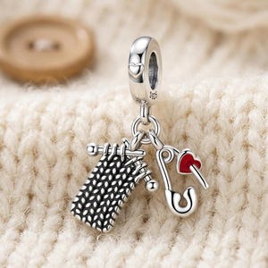 Op de afbeelding: Een bedel van sterling zilver met een gebreid vierkantje, twee breinaalden en een veiligheidsspeld met een klein rood hartje. Het oogje is gestempeld met 925 en voorzien van een klein hartvormig detail voor aan een armband.